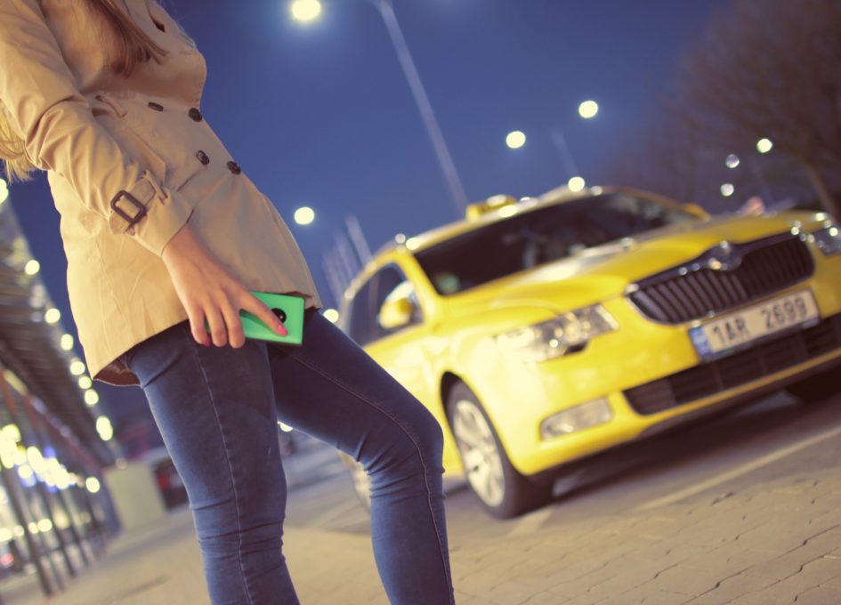 A woman holding a cell phone waits for a taxi to take her where she needs to go that night.